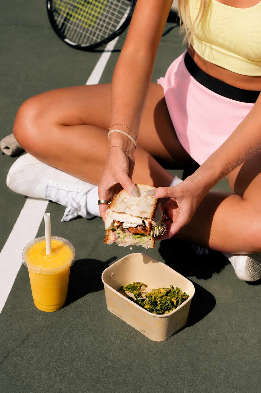 Woman sitting courtside on a tennis court with a sandwich, greens and a mango smoothie