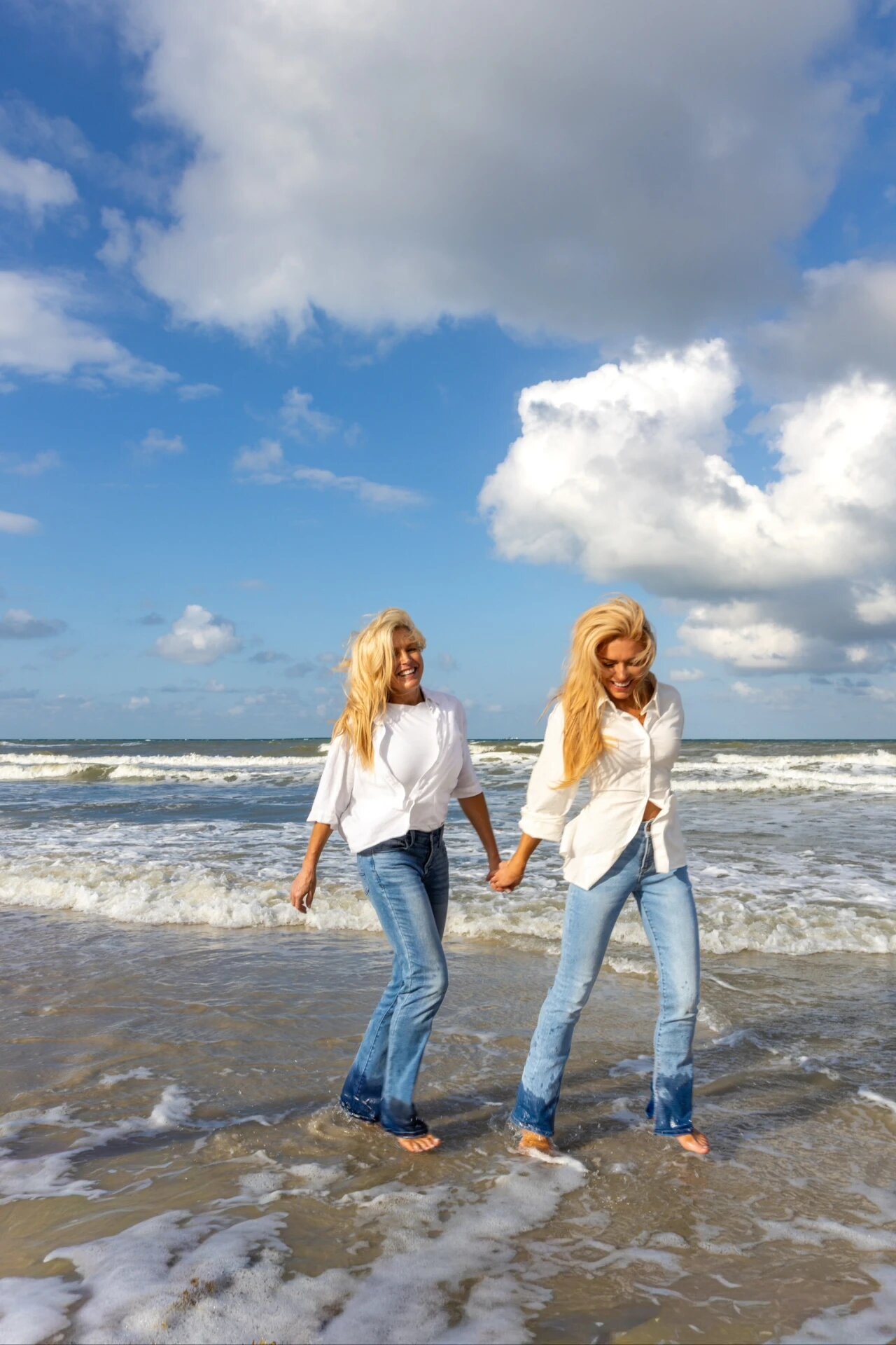 Two smiling women in white shirts and jeans holding hands while wading through the surf