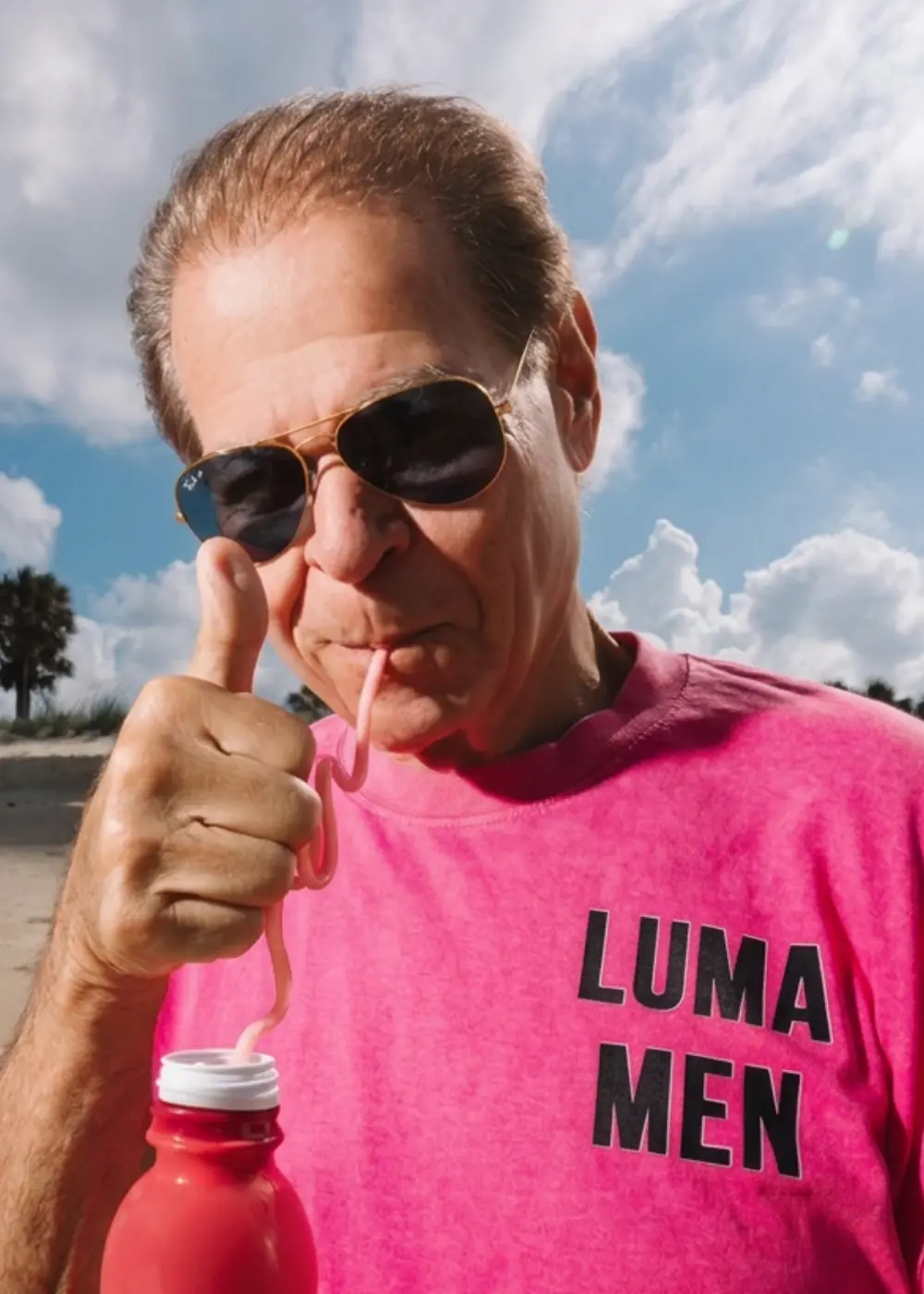 Man in a pink LUMA MEN tee giving a thumbs up with a drink at the beach